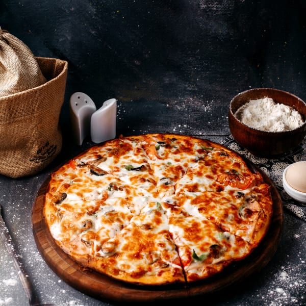 A sliced mushroom and cheese pizza on a wooden board, with flour and ingredients scattered around.