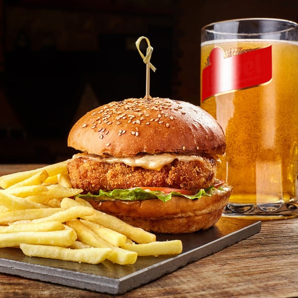 Crispy chicken sandwich with fries and a glass of beer on a dark wooden table.